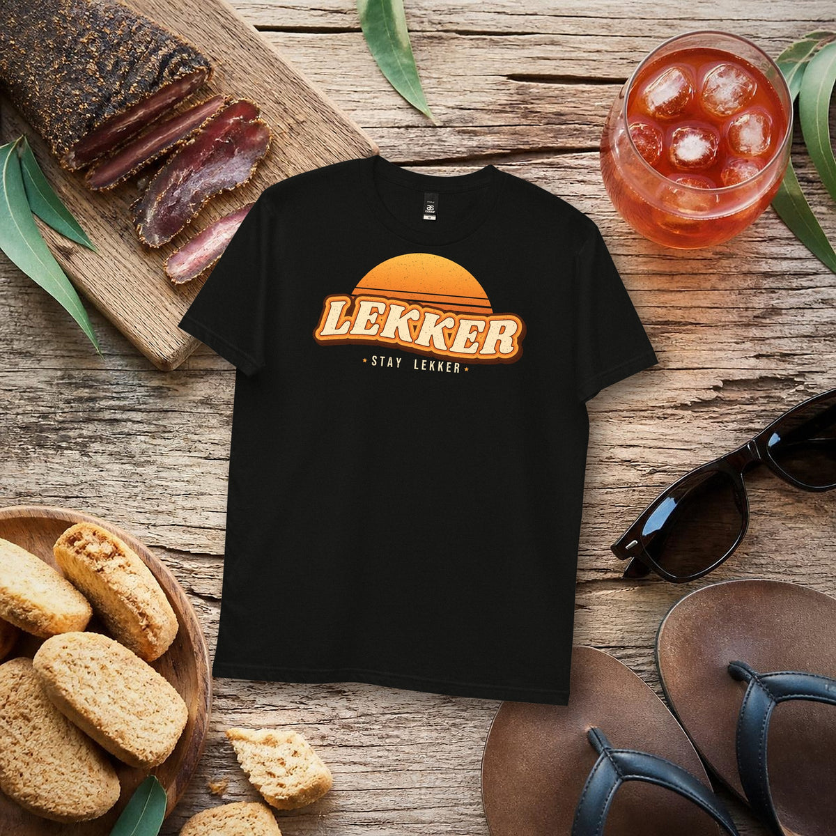 Stay Lekker Tee in black with retro sunset 'LEKKER – stay lekker' print, flat lay with biltong, rusks and rooibos