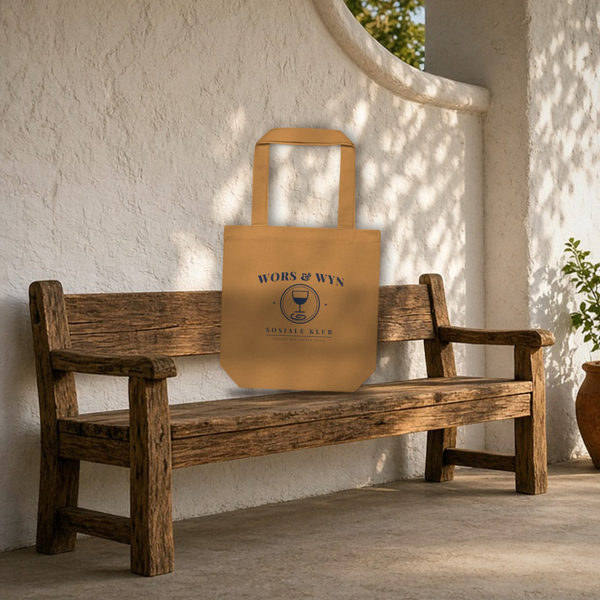 Wors & Wyn Sosiale Klub Tote Bag in camel leaning on a wooden bench on a Cape Winelands patio with a whitewashed gable wall