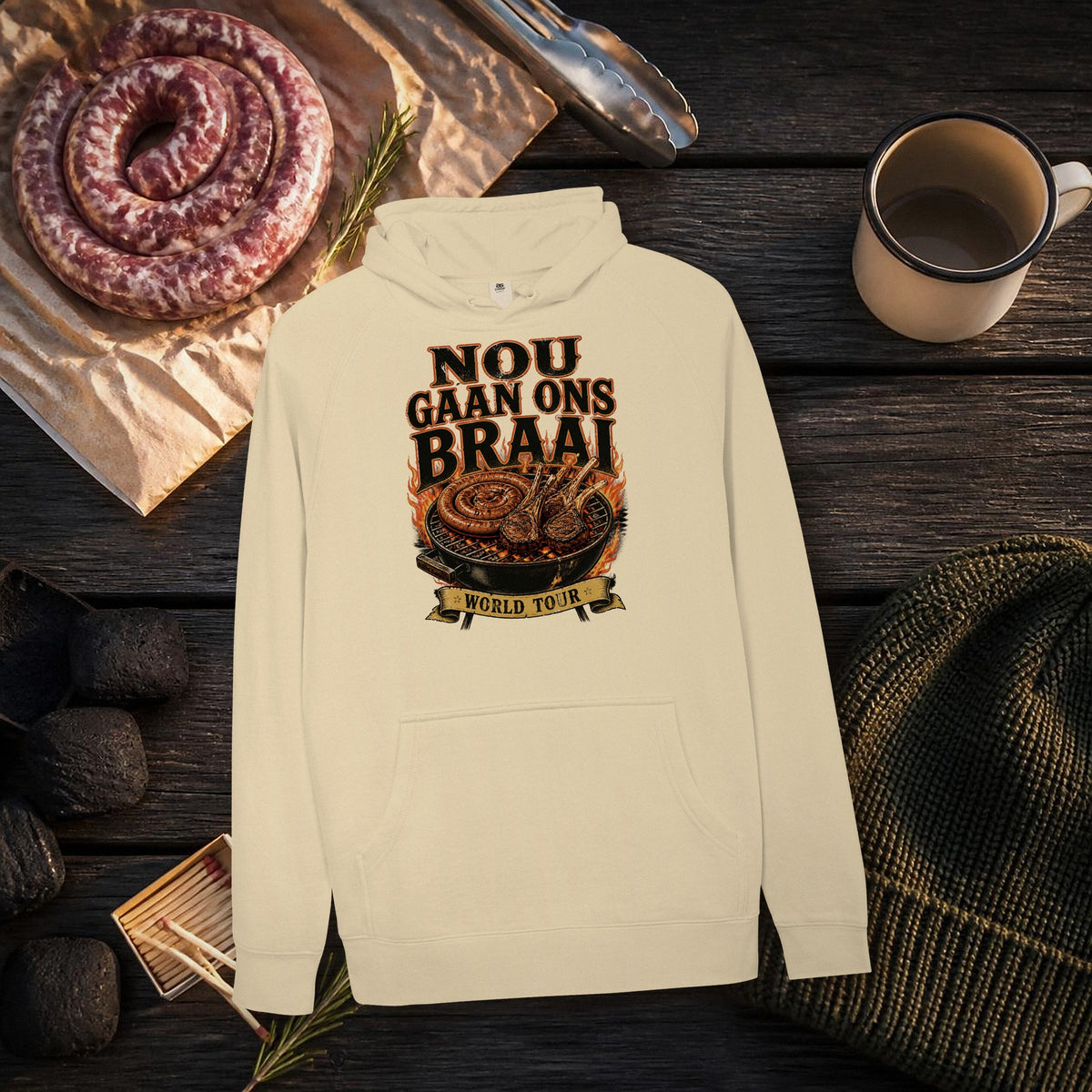 Nou Gaan Ons Braai Hoodie flat lay with boerewors, charcoal and a beanie – 'World Tour' braai grill print