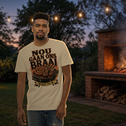 Nou Gaan Ons Braai Tee in natural – man wearing the 'Nou Gaan Ons Braai World Tour' print t-shirt by a backyard brick braai at dusk