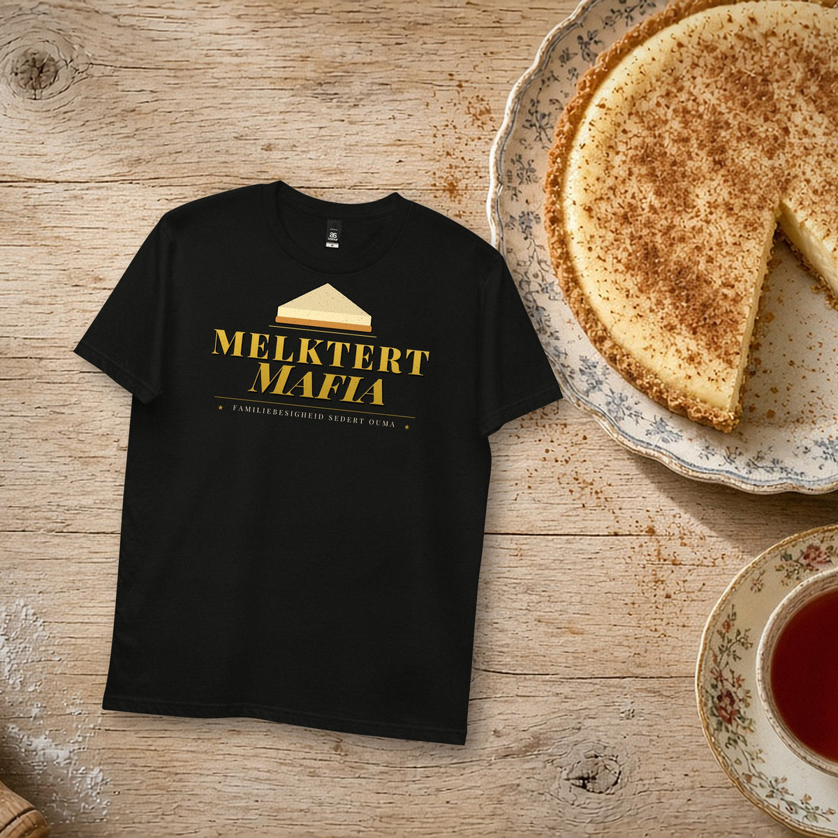 Melktert Mafia Tee in black – flat lay with a homemade melktert, rooibos tea and a rolling pin, gold 'Melktert Mafia' print